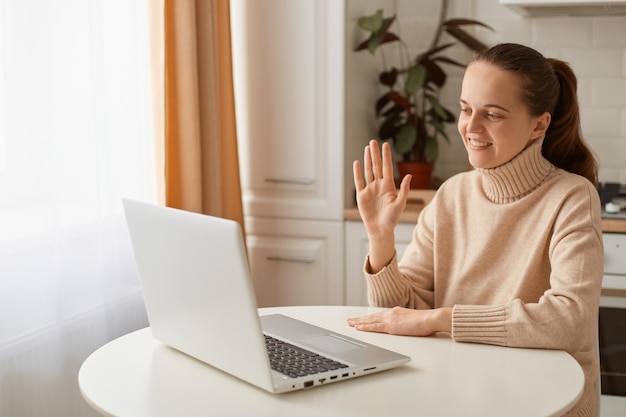 Indoor shot of Caucasian young adult beautiful woman with ponytail hairstyle wearing casual style beige sweater sitting at table in kitchen and having video call via laptop, waving hand, saying hello.