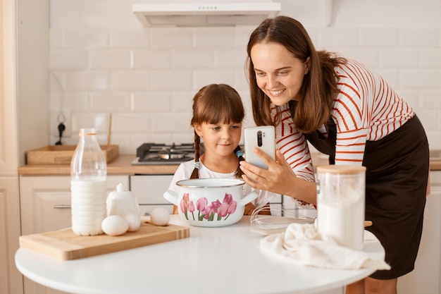 Indoor shot of Caucasian young adult woman cooking with her daughter in the kitchen, browsing internet for finding recipe, looking smiling at device, being happy to bake together.