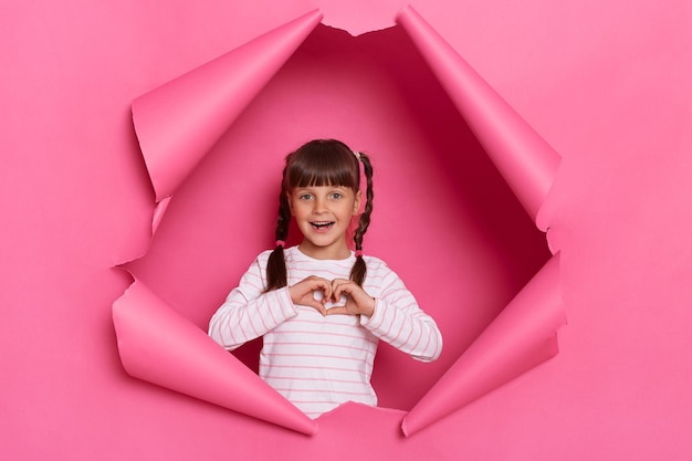 Ripresa in interni di affascinante bambina eccitata che indossa una camicia a righe in posa in carta rosa strappata in piedi guardando la fotocamera che mostra il gesto del cuore con i palmi delle mani