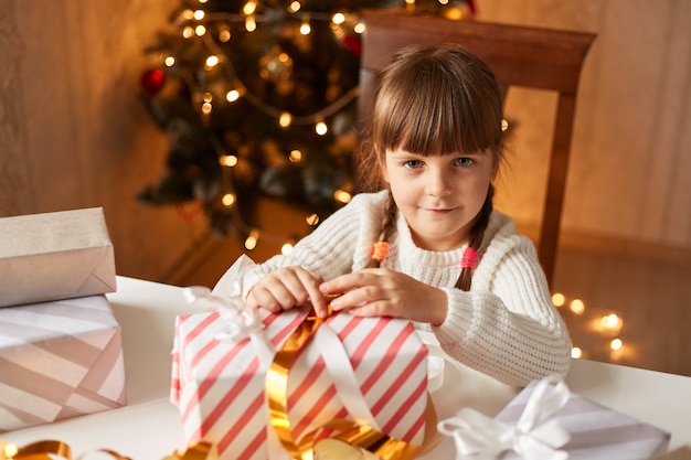 Крытый снимок очаровательного позитивного очаровательного ребенка женского пола в белом свитере, смотрящего в камеру, упаковывая рождественские подарки, празднуя канун Нового года.