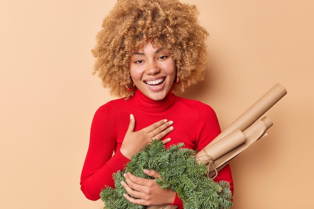 Indoor shot of cheerful curly woman makes thankful gesture smiles gladfully feels happy holds green wreath and paper for wrapping presents wears red turtleneck isolated over beige background