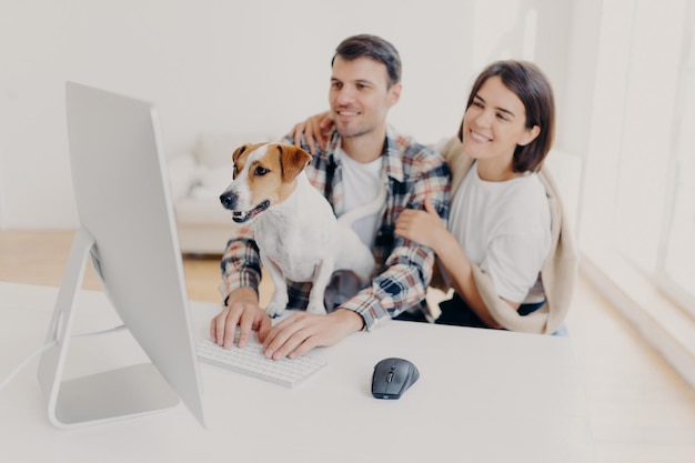 Indoor shot of cheerful husband and wife look with joyful expressions laugh as watch funny movie rest together at free time curious dog looks attentively at monitor of computer man keyboards