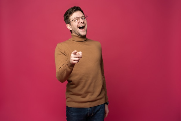 Indoor shot of cheerful man in spectacles and casual clothes indicates happily at you