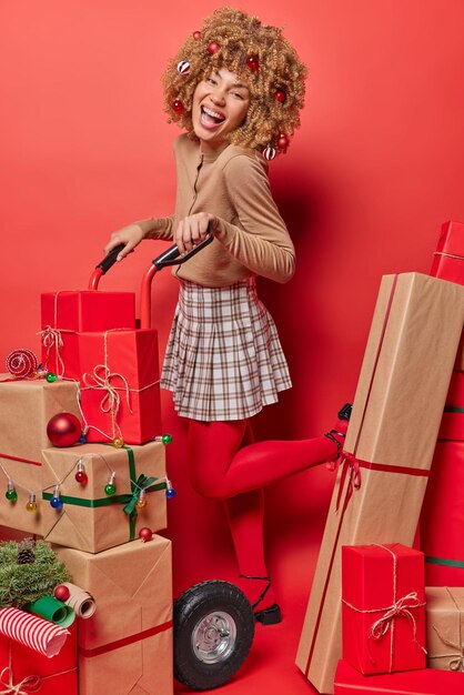 Indoor shot of cheerful woman with curly hair carries track\
with stack of gift boxes laughs joyfully celebrates new year at\
home prepares presents for everybody busy with holiday\
preparations