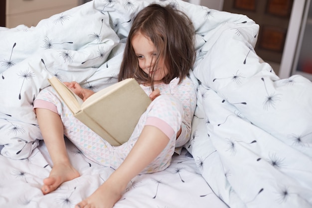 Indoor shot of child reading book in bed under blanket with dandelion. Female kid spending time in cozy bedroom. Little girl doing homework before sleepping, reading fairytale. Childhood concept.