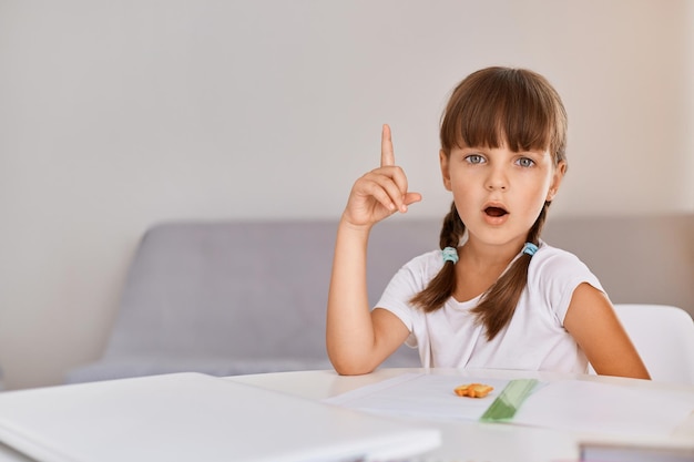 Indoor shot of clever smart child girl doing homework writing and reading at home sitting at table looking at camera raising finger up having great idea