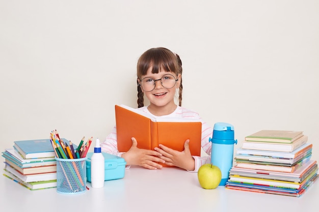 Indoor shot of clever smiling cute little girl with braids sitting at desk holding book reading interesting text enjoying studying process posing isolated over white background