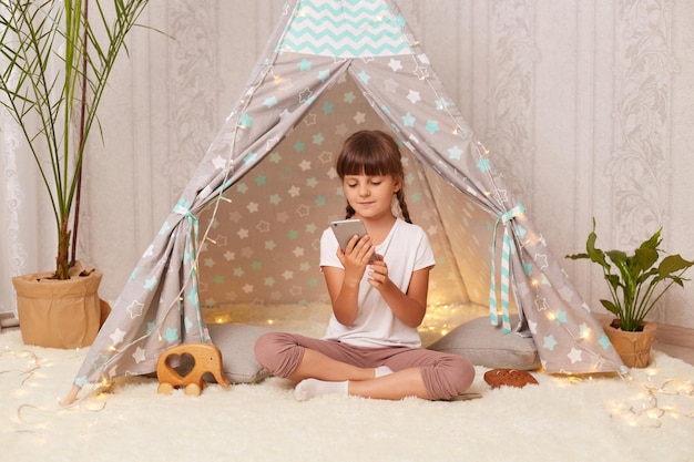 Indoor shot of concentrated little girl with braids wearing white t shirt posing in wigwam cute kid holding mobile phone and checking social networks