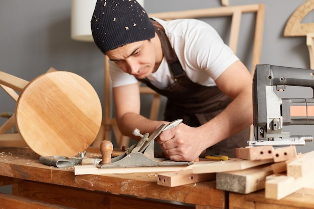 Indoor shot of concentrated young carpenter wearing apron and cap working in workshop using plane making wooden chair enjoying his work looking at plank with serious expression