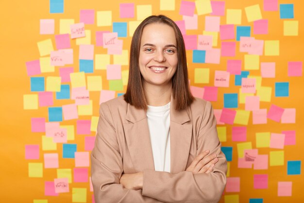 Indoor shot of confident businesswoman wearing beige jacket\
standing against yellow wall covered with stickers keeps hands\
crossed looking at camera with smile expressing confidence