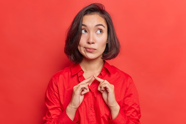 Indoor shot of contemplative Asian female model keeps index fingers together tries to gather with thoughts considers something focused above dressed casually isolated on vivid red 