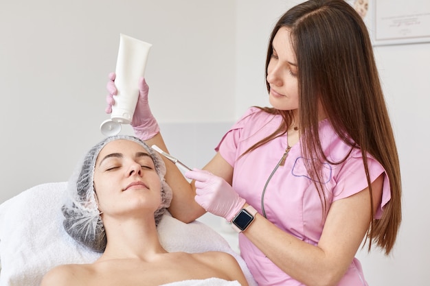 Indoor shot of cosmetologist applying to her client anti ageing mask with brush, holds big bottle of cream in hands. lady keeps eyes closed, ralaxes during moisturizing and rejuvenation procedures.