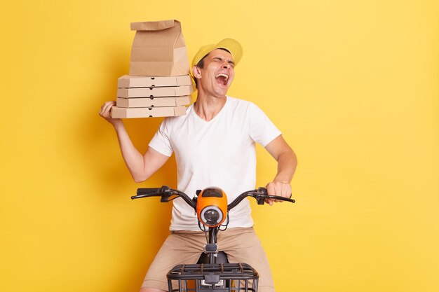 Indoor shot of crazy silly overjoyed delivery man with pizza in cardboard box sitting on fast motorbike isolated over yellow background laughing out loud