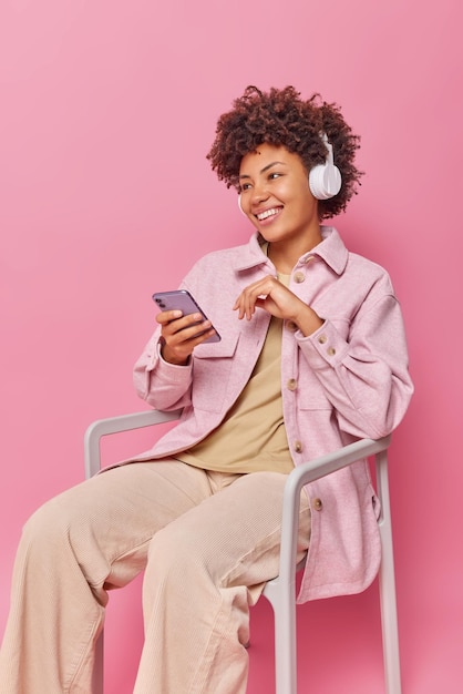 Indoor shot of curly haired female student learns information from audio uses wireless headphones and smartphone poses on chair looks away isolated over pink background Curly woman enjoys music