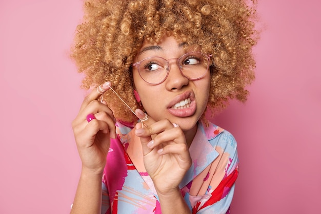 Indoor shot of curly haired young woman holds dental floss\
purses lips looks away wears spectacles and colorful shirt isolated\
over pink background oral care and daily hygiene routine\
concept