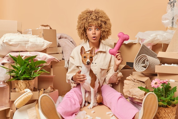 Indoor shot of curly haired young woman poses with pet dressed\
in jacket trousers and sportshoes going to relocate to new house\
packs things in cardboard boxes isolated over beige background