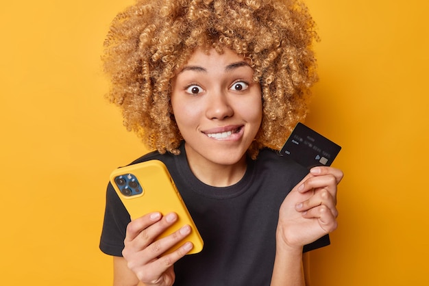 Снимок в помещении: кудрявая женщина кусает губы, удивленно смотрит в камеру, держит смартфон и кредитную карту для оплаты онлайн, носит повседневную черную футболку, позирует на желтой стене, делает покупки в Интернете