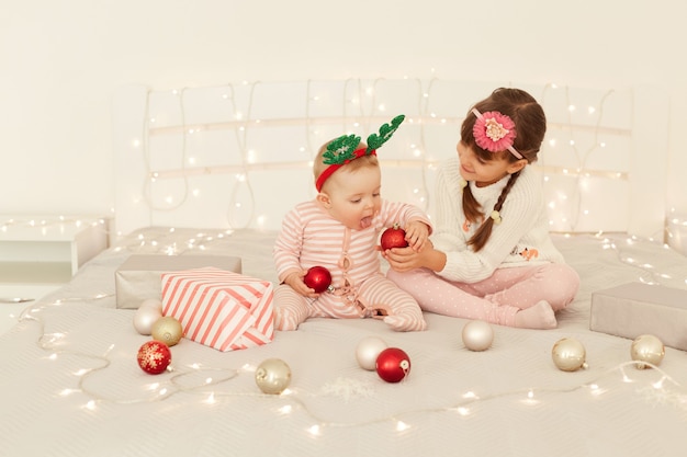 Indoor shot of cute charming little girls wearing casual clothing and party hoops sitting party on bed, elder sister giving red baubles to her sister, celebrating New year holidays.