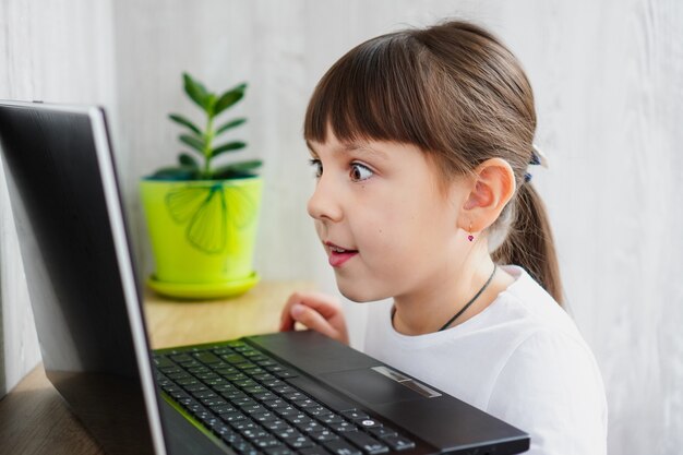 Indoor shot of cute dark haired female child looking surprised at notebook