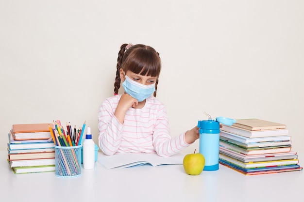 Indoor shot of cute sick unhealthy little schoolgirl wearing protective mask during visiting school kid with dark hair and braids sitting at table surrounded with books touching water bottle