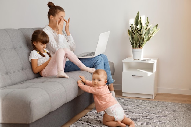 Indoor shot of cute toddler baby crawling to mother wearing shirt and jeans sitting on sofa with elder daughter, working on laptop, freelancer doing her work, being tire, feels eyes hurt, rubbing eye.