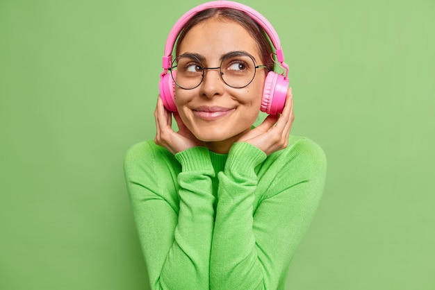 Indoor shot of dreamy satisfied woman keeps hands on pink streo headphones listens music with pleased thoughtful expresssion wears round spectacles and jumper isolated over vivid green background