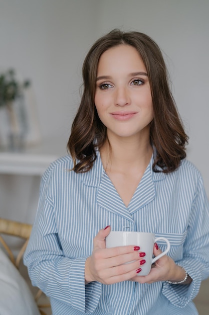 Indoor shot of dreamy young European woman with charming look dressed in striped pyjamas drinks coffee thinks about something enjoys calm domestic atmosphere People drinking and home concept