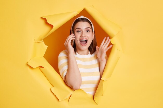 Indoor shot of excited amazed woman wearing striped t shirt looking through breakthrough of yellow background talking phone with her friend gossiping