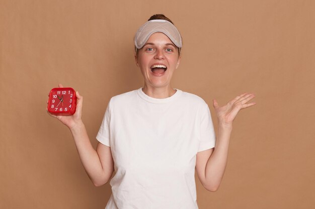 Indoor shot of excited amazed woman wearing white t shirt and blindfold holding red alarm clock wake up early with good mood posing isolated over brown background