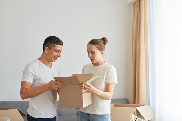 Indoor shot of excited couple standing with cardboard package in the new apartment looking inside box with open mouths expressing astonishment wearing white casual t shirts