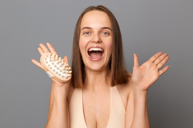 Indoor shot of excited joyful woman uses dry body brush wants to have smooth clean skin yelling with excitement poses against gray background posing with open mouth