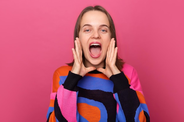 Indoor shot of excited woman wearing sweater posing against pink wall screaming loud with hands near mouth making announcement saying important information