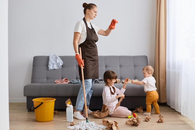 Indoor shot of exhausted sad upset woman with bun hairstyle wearing brown apron looking at mess on the floor after cleaning room looks at her children sitting on floor and throwing paper