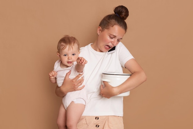 Indoor shot of exhausted shocked woman wearing white casual style shirt holding in arms toddler daughter looks at the spoiled food in the pot while talking on smartphone isolated on brown background