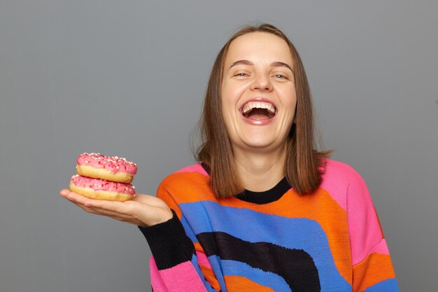 Indoor shot of extremely happy woman with brown hair standing\
holding donuts in hand laughing being in good mood looking at\
camera posing isolated over gray background