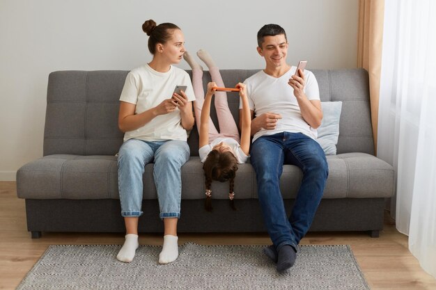 Indoor shot of family sitting on sofa together father showing shock content and laughing woman looking with open mouth and surprised expression gadget addiction