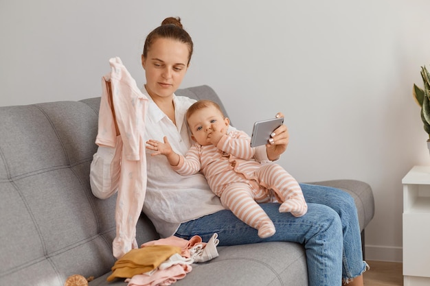 Indoor shot of female blogger with bun hairstyle wearing white shirt and jeans sitting on cough with her toddler kid and holding smart phone and daughter's new clothing in hands.