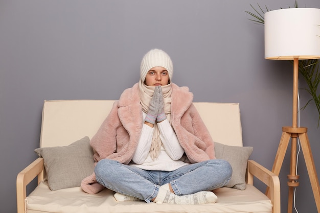 중앙 난방 없이 아파트에서 따뜻한 생활을 하려고 집에서 소파에 앉아 모자와 겨울 코트를 입은 얼어붙은 매력적인 여성의 실내 사진