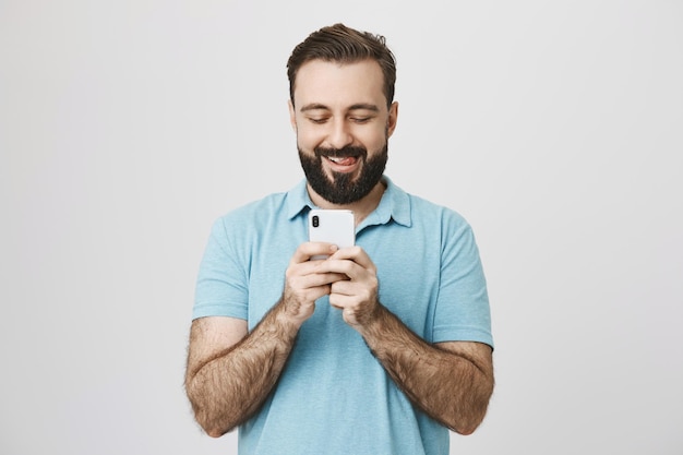 Indoor shot of funny man playing games on smartphone or messaging holding it with both hands smiling and sticking out tongue over gray wall