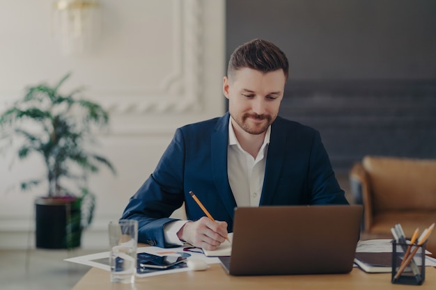 Indoor shot of handsome male office worker rewrites information from notepad in diary sits at desk dressed in formal clothes spends time for searching info. Businessman takes notes does paper work