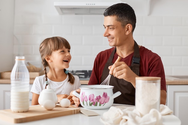 Indoor shot of handsome man sitting at table with daughter and cooking together in kitchen preparing dough for cake looking at each other with smile