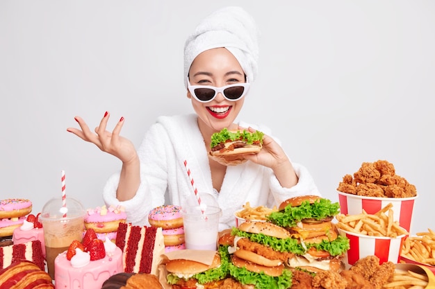 Indoor shot of happy Asian woman raises hand smiles sincerely holds tasty hamburger 