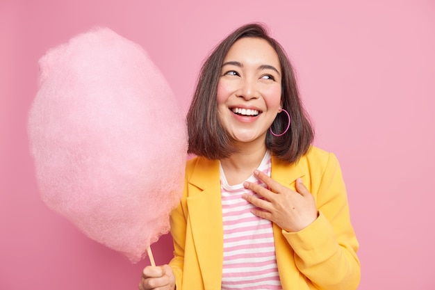 Indoor shot of happy Asian woman with dark hair smiles broadly recalls something funny enjoys spare time holds appetizing sweet cotton candy focused aside wears yellow jacket isolated on pink