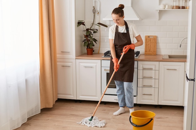 Indoor shot of happy Caucasian woman housewife cleaning floor with mop in kitchen wearing white t shirt jeans and brown apron expressing positive emotions while washing her flat