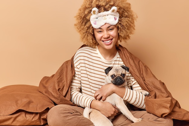 Indoor shot of happy curly haired pet owner wears pajama and sleepmask stays in bed with favorite pet isolated over brown background European woman enjoys rest together with domestic animal