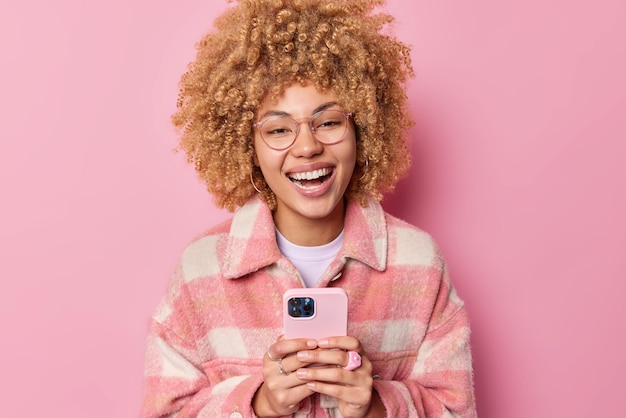 Indoor shot of happy curly haired woman laughs joyfully dressed\
in casual checkered jacket uses mobile phone connected to wifi\
types feedback enjoys cellphone messaging isolated over pink\
background