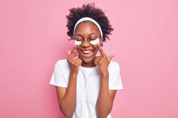 Indoor shot of happy dark skinned young woman with curly hair points at beauty patches under eyes smiles broadly 