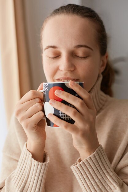 目を閉じて飲み物を楽しんで、カップからホットコーヒーやお茶を飲むベージュのカジュアルスタイルのセーターを着ているポニーテールの髪型で幸せな喜びの満足している女性の屋内ショット。