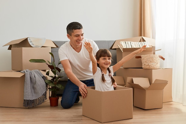 Indoor shot of happy family having fun during moving in a new apartment, expressing positive emotions, little girl sit in a cardboard box with raised arms, relocation, playing with father.