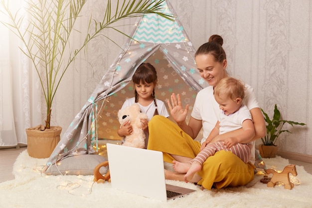 Photo indoor shot of happy family mother elder daughter and toddler baby sitting on floor near peetee tent and using laptop for talking on video call waving hand saying hello